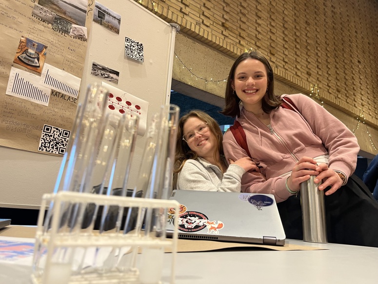 To smilende piger står tæt sammen bag et bord i et udstillingslokale. Den ene har briller på, og den anden bærer en lyserød hættetrøje og holder en termoflaske. I forgrunden ses et stativ med reagensglas.