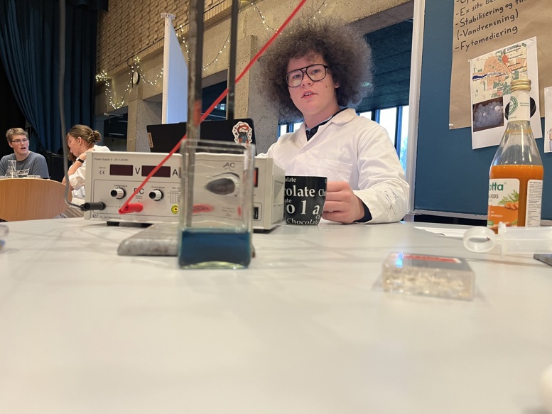 En ung mand med stort mørkt krøllet hår og briller sidder bag en videnskabelig testopstilling. Han bærer en hvid kittel og holder en kop, mens han kigger direkte mod kameraet. I forgrunden ses en lille beholder med blå væske.