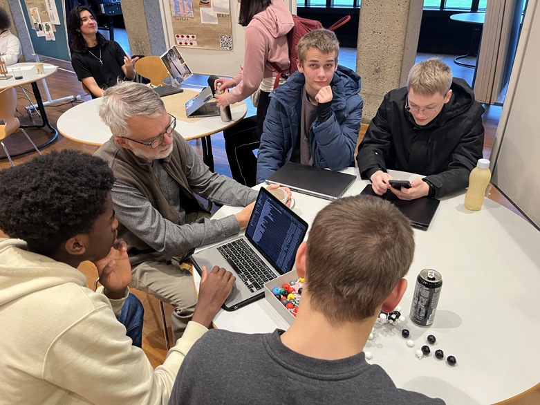 En ældre mand med briller og skæg vejleder tre unge drenge ved et bord. De kigger på en bærbar computer, der viser rækker af kode, mens der ligger molekylmodeller spredt på bordet.