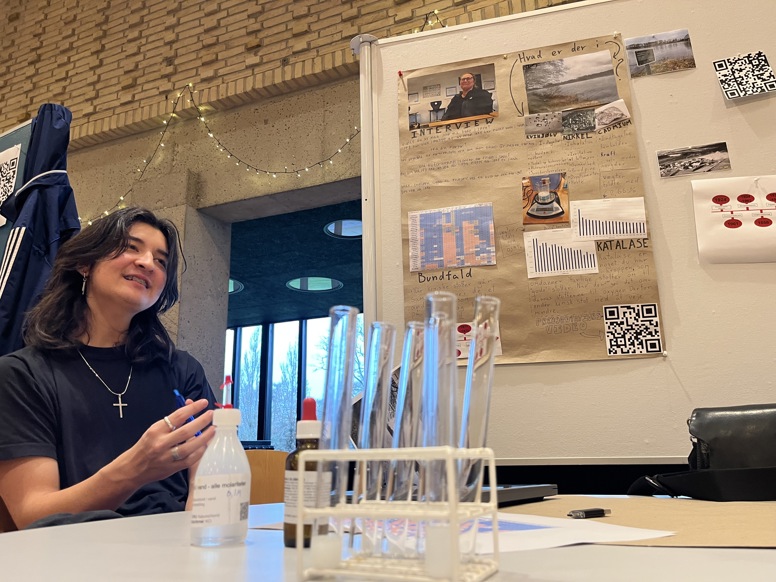 En ung person med mørkt hår og et kors-halskæde sidder ved et bord og forklarer noget. I forgrunden ses reagensglas i et stativ, og i baggrunden hænger en planche med titlen "Hvad er der i?" og grafer over kemiske stoffer.