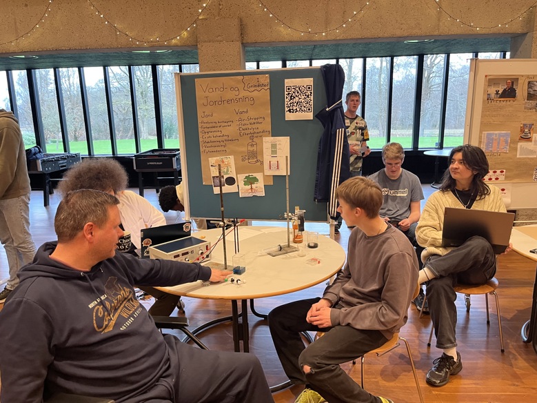 En mand i en mørkeblå hættetrøje sidder ved et bord og peger på en videnskabelig opstilling. Ved siden af ham sidder en ung elev og lytter. I baggrunden ses en planche om rensning af jord og vand.